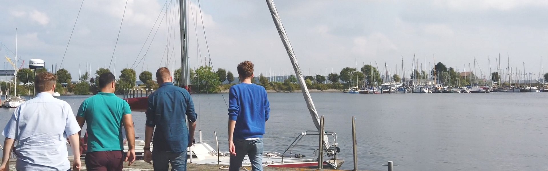 Vier GMSH-Studenten gehen am Kieler Yachthafen entlang. Im Hintergrund ist Wasser und ein Segelboot zu sehen. 