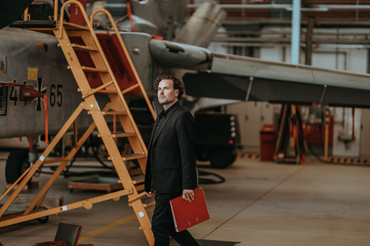 GMSH-Projektleiter Thomas Friedrich steht in einer Halle, im Hintergrund steht ein Kampfjet.